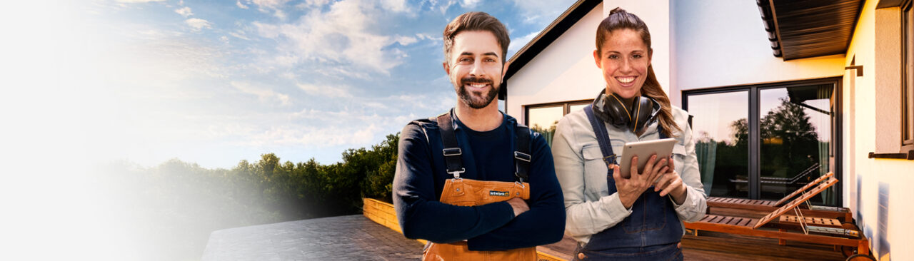 Ein Handwerker und eine Handwerkering stehen vor einem modernen Neubau mit großer Terrasse und Garten